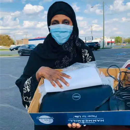 The photo shows a adult woman wearing a blue mask. She is a CORE student picking up a donated laptop to continue her classes virtually. There is a blue sky and white clouds behind her.